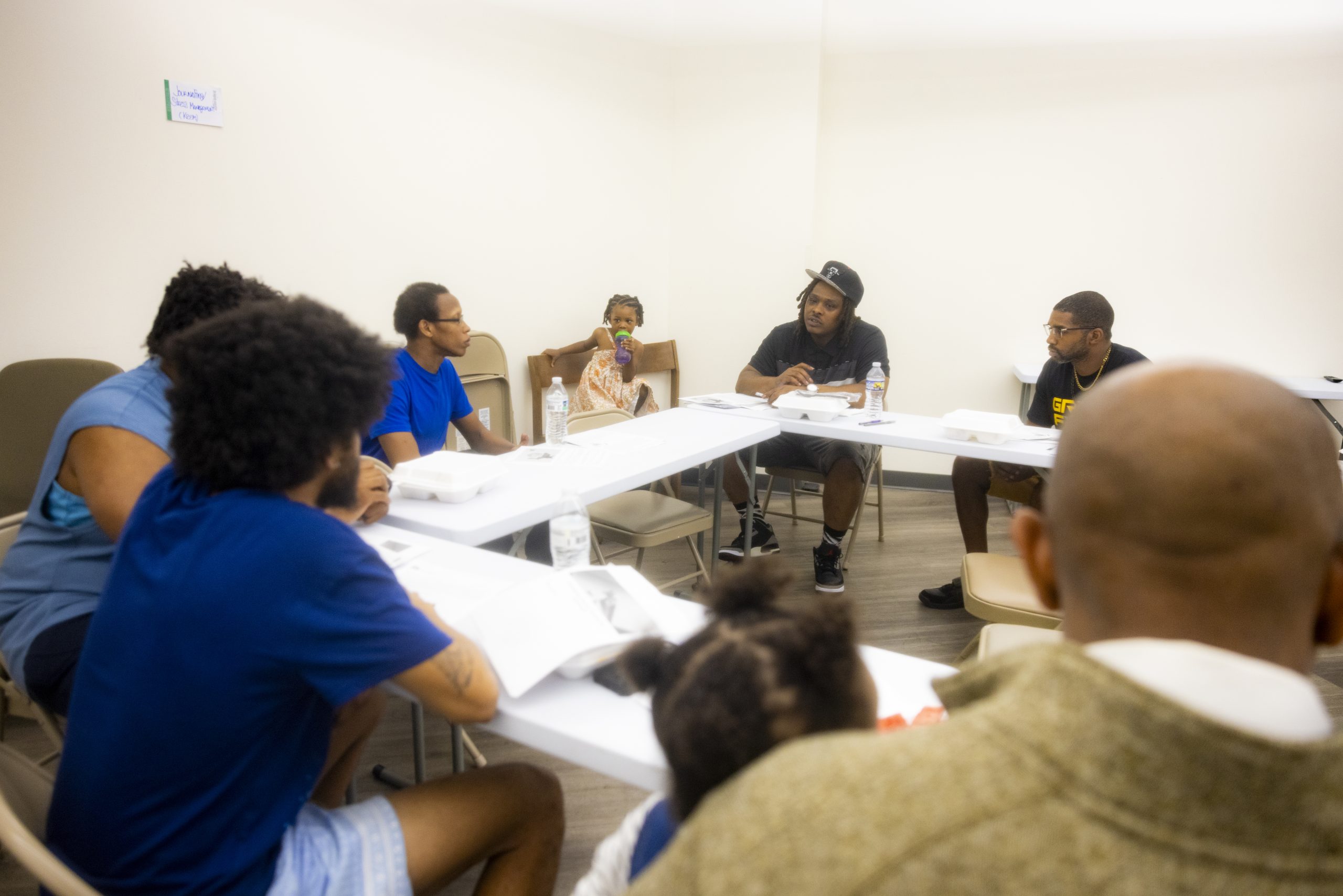 A group of people sit around tables engaged in discussion, with papers, water bottles, and food containers in front of them in a brightly lit room.