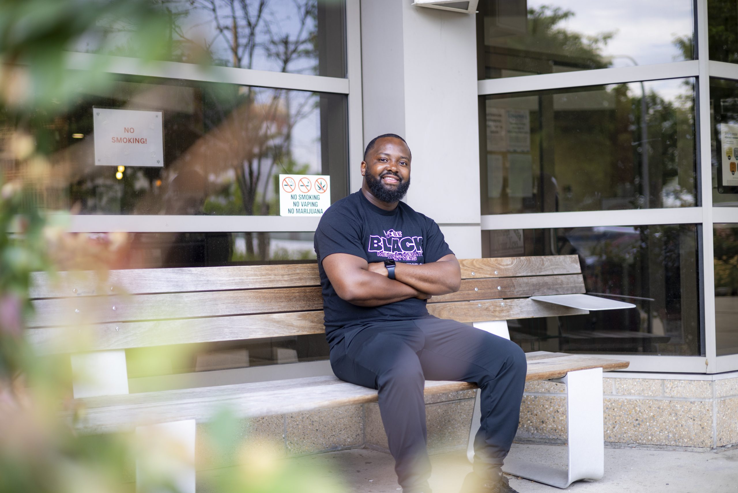 A man sits on a wooden bench outside a building with glass windows, arms crossed, looking at the camera. There are