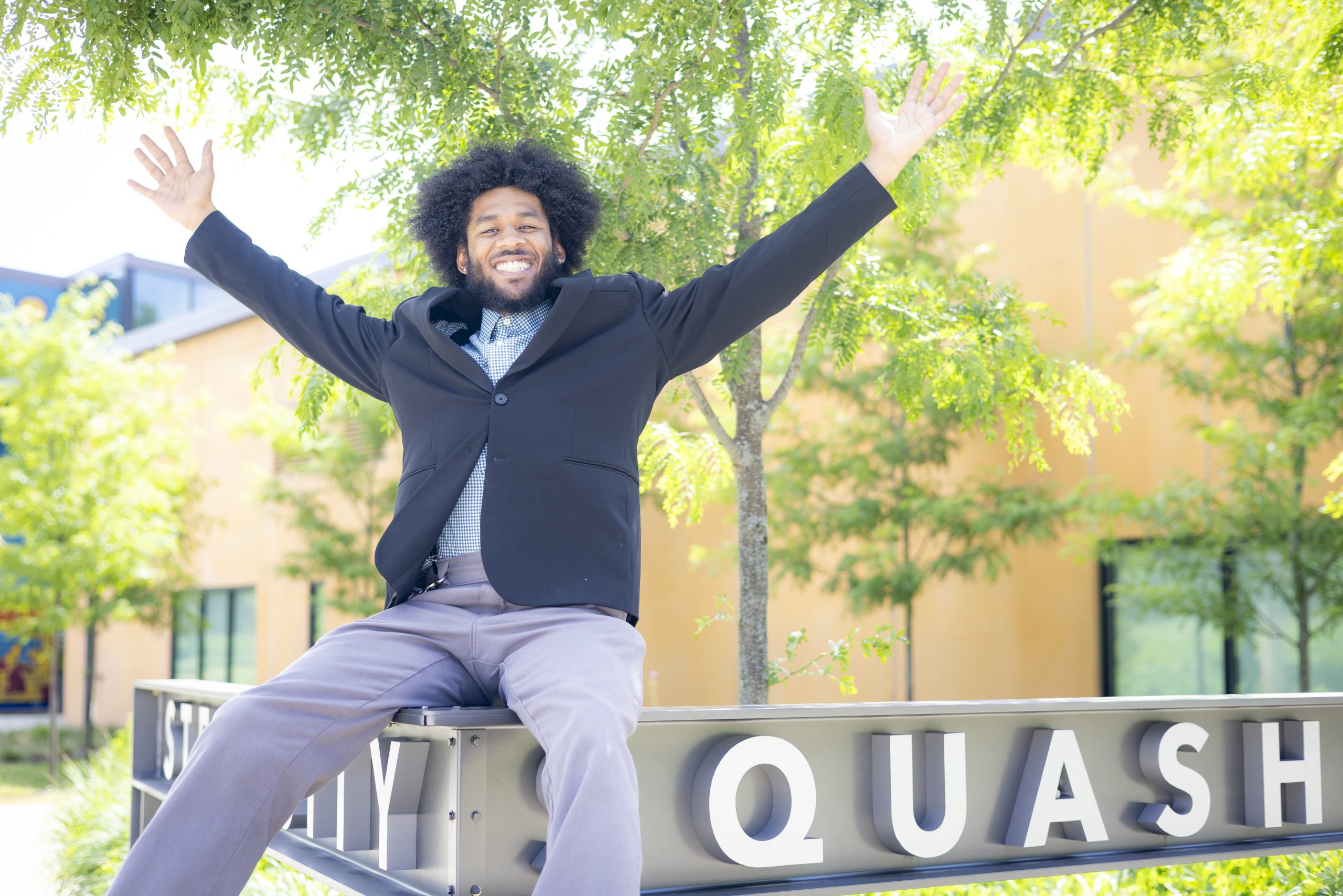 A man in a suit sits on a metal sign with his arms raised and smiling, outdoors on a sunny day with trees and a building in the background.
