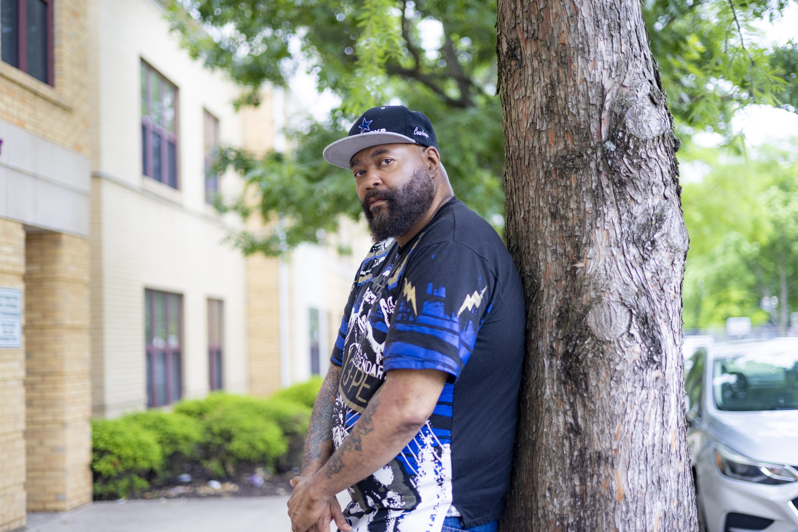 A man wearing a cap and graphic t-shirt leans against a tree on a city sidewalk, with buildings, shrubs, and parked cars in the background.