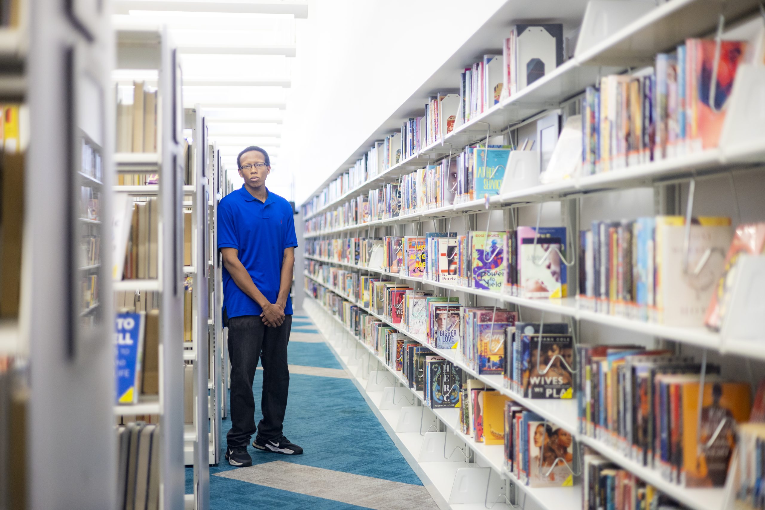 A person in a blue shirt stands in the aisle of a library beside shelves filled with books.