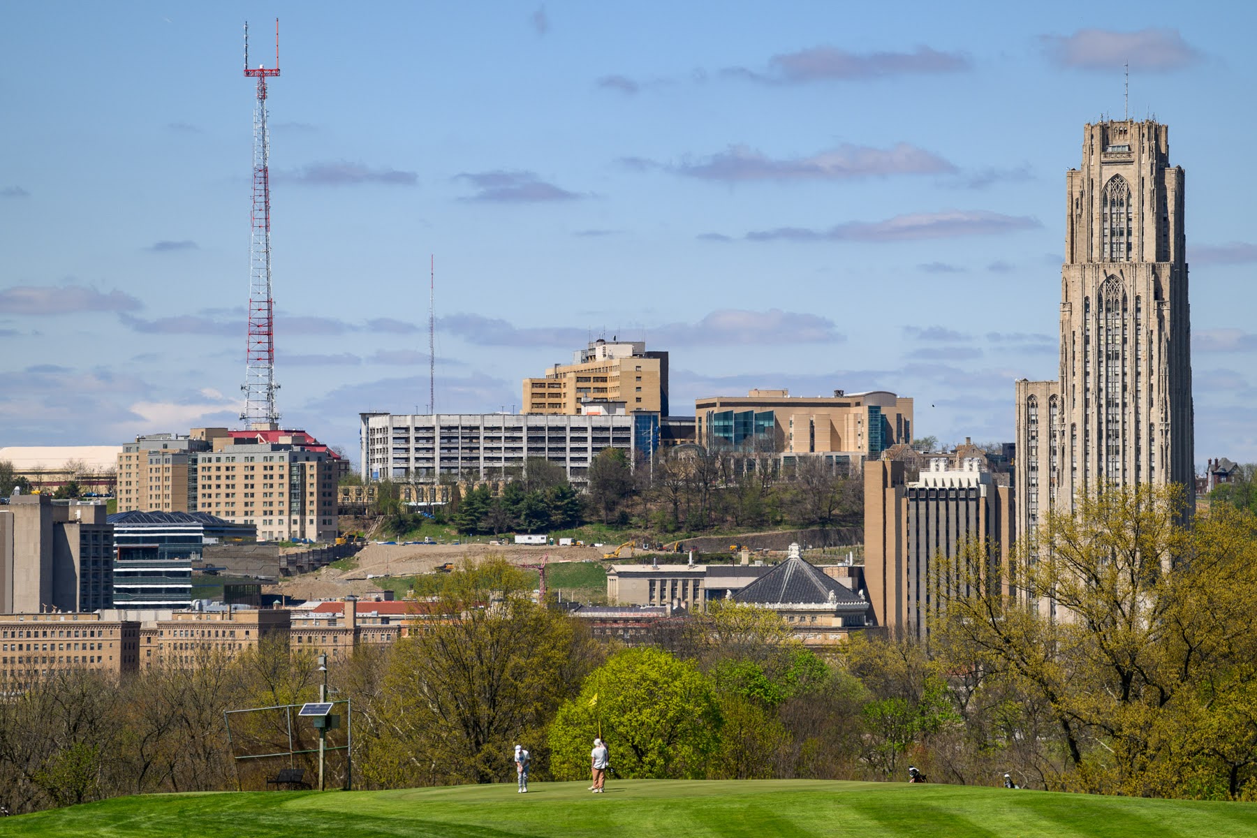 Distant view of a city skyline with tall buildings, radio towers, trees, and two people walking on a grassy area in the foreground under a partly cloudy sky.