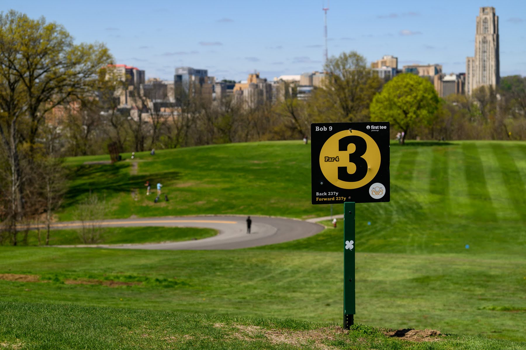 A golf course with a sign marking hole 3, par 3, with a city skyline and trees visible in the background.