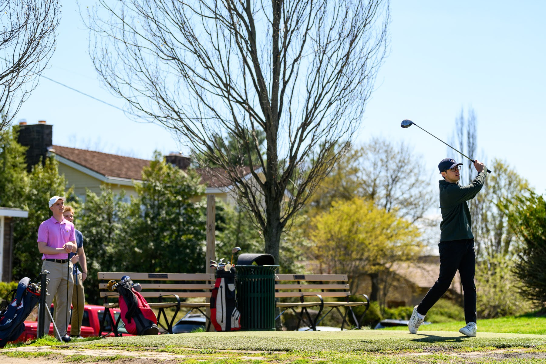 Two people at a golf course; one person swings a golf club while the other watches. Golf bags and benches are nearby, with trees and houses in the background.