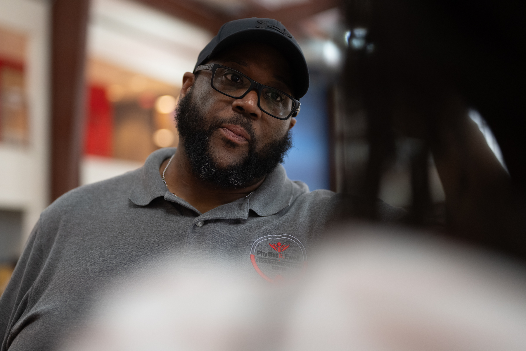 A man in a gray polo shirt and black cap looks attentively at someone off-camera in an indoor setting.