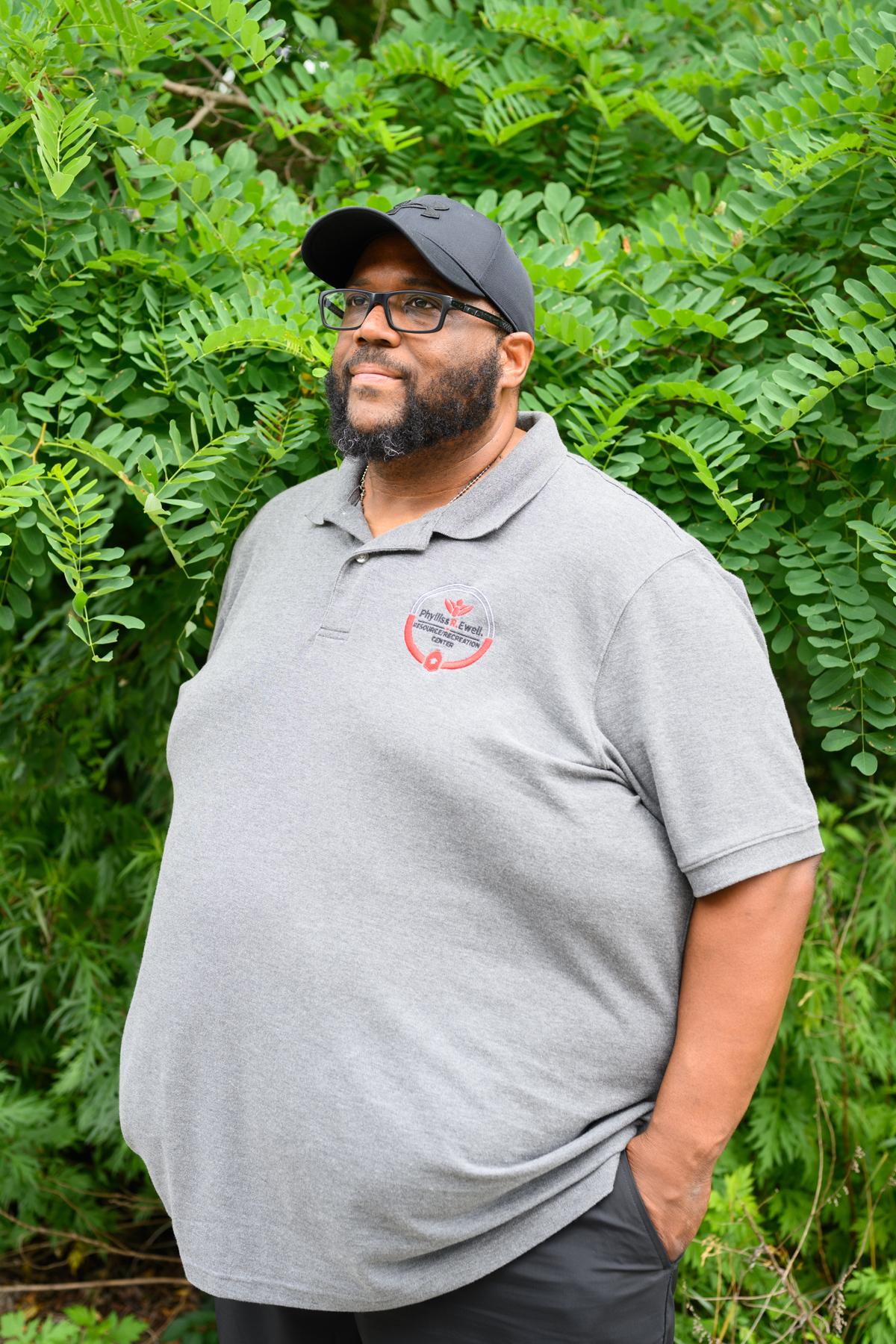 A man wearing glasses, a black cap, and a gray polo shirt stands outdoors in front of green foliage with his hand in his pocket.