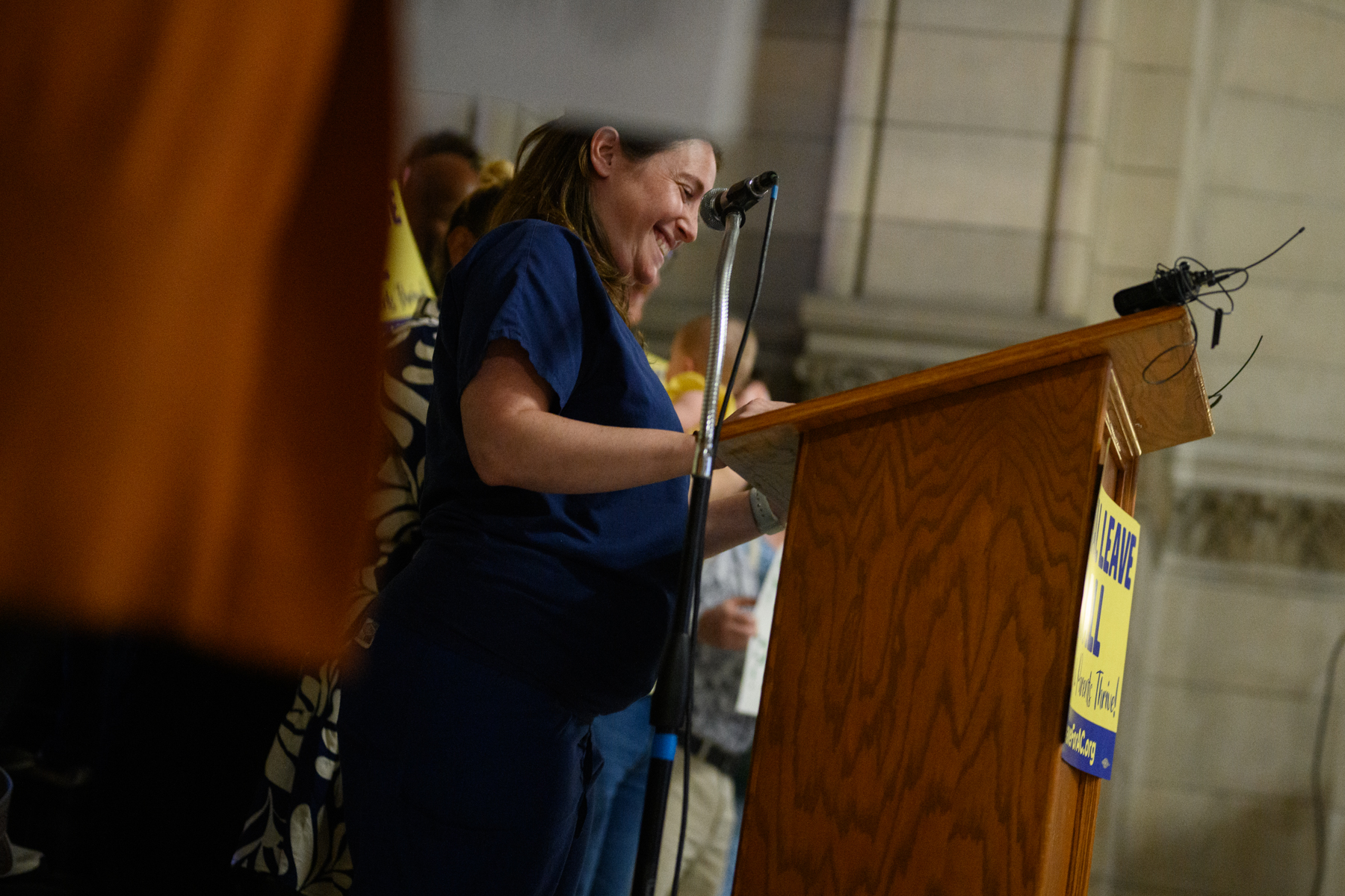 A woman stands at a podium, speaking into a microphone at an indoor event, with people gathered in the background.
