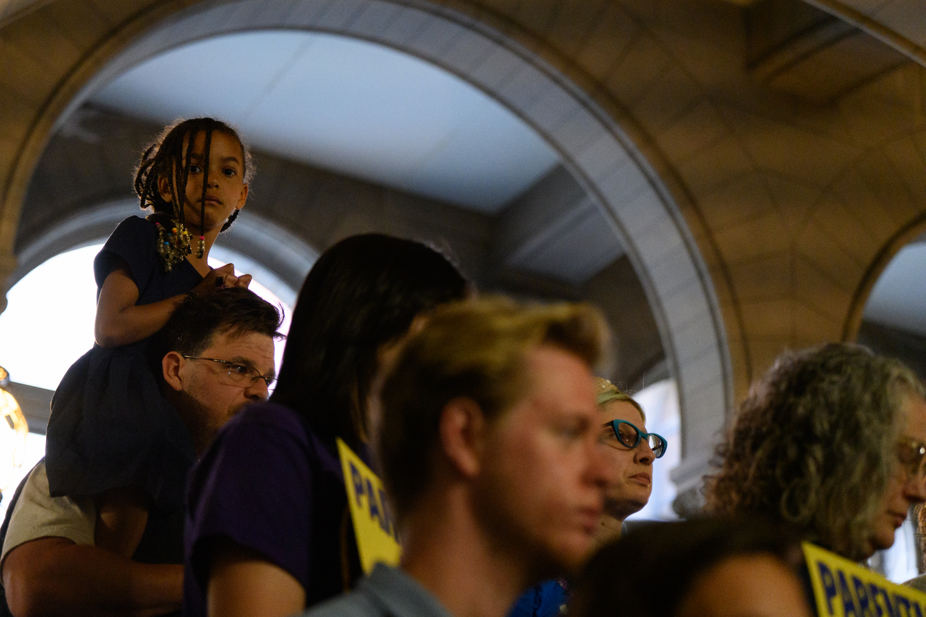 A young girl sits on an adult's shoulders among a group of people indoors, some holding yellow signs.