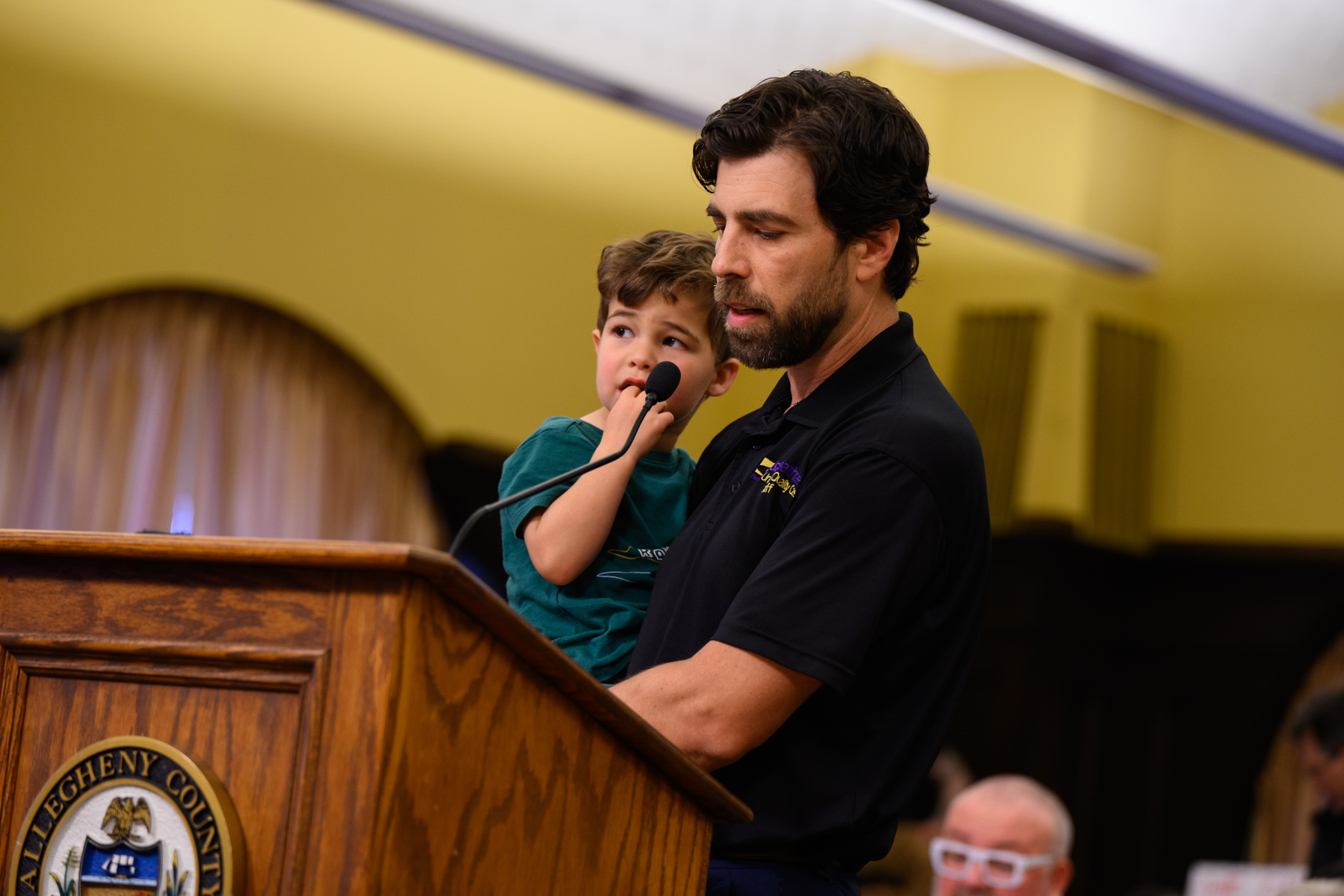 A man holds a young boy at a wooden podium with a microphone in an indoor setting, speaking to an audience.