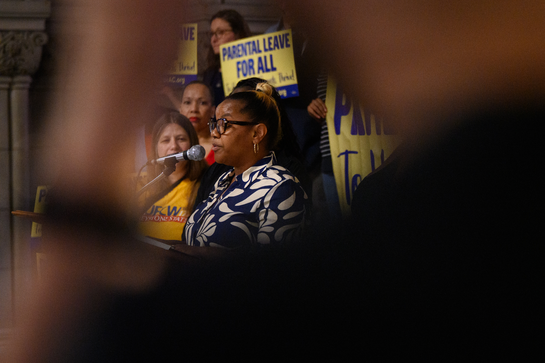 A woman speaks at a microphone during a rally, with people behind her holding signs that read