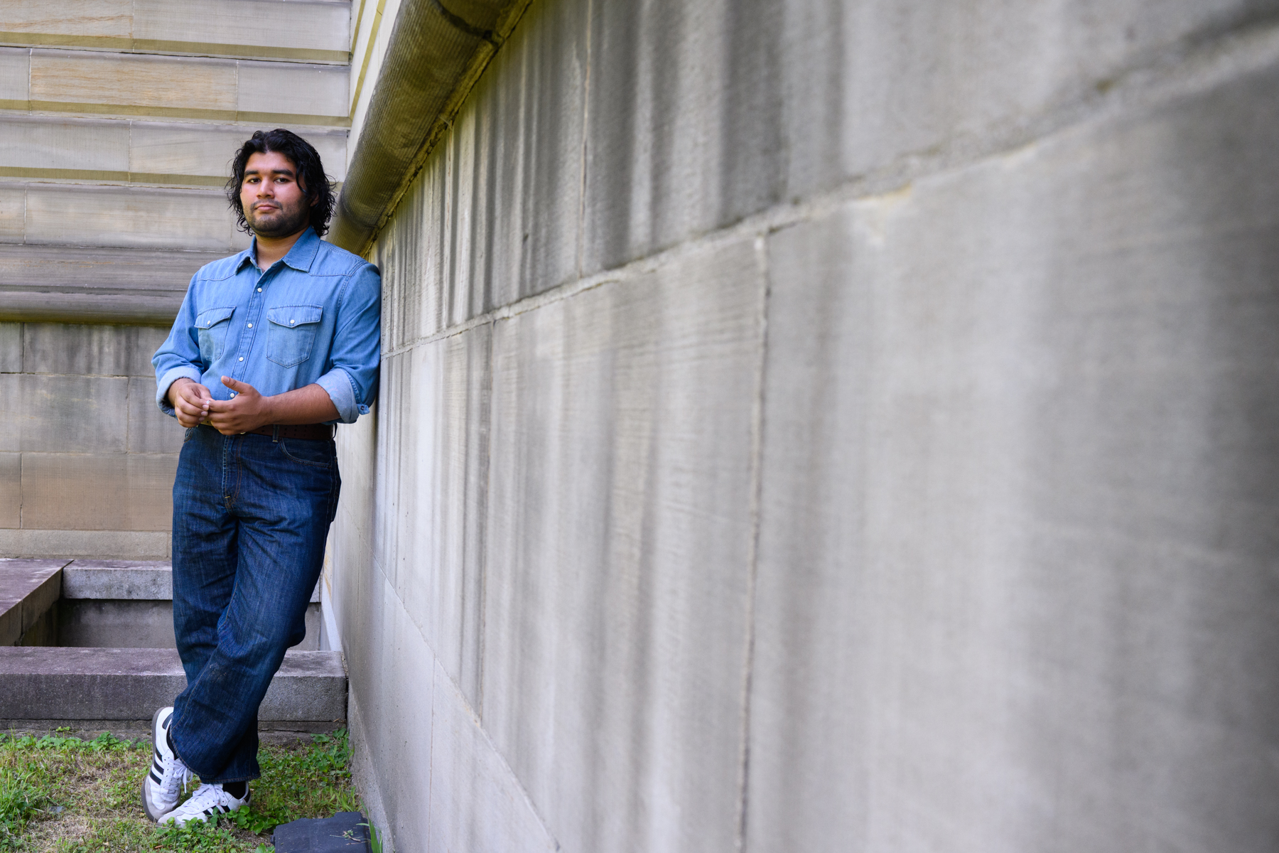 A man in a blue denim shirt and jeans stands leaning against an exterior stone wall, holding a phone and looking at the camera.