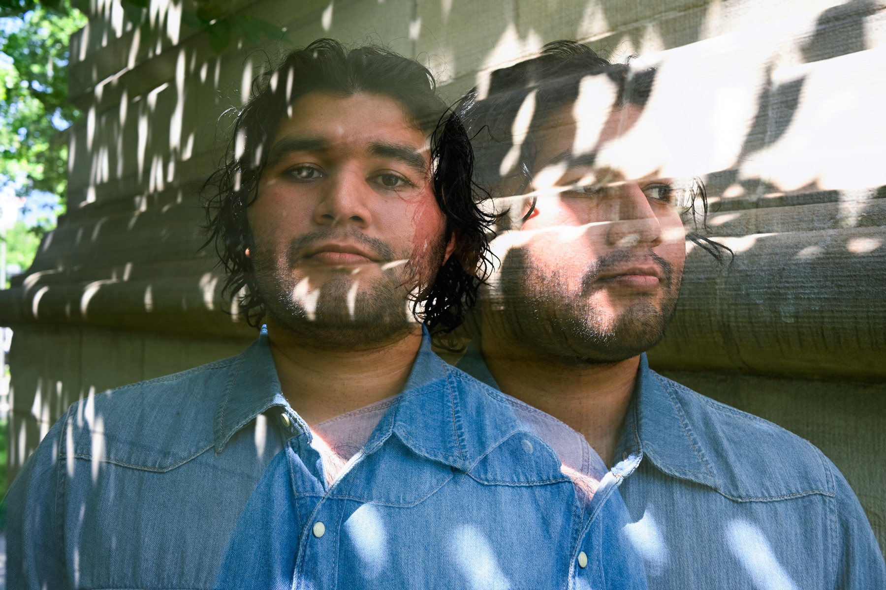 A person with dark hair and a denim shirt appears twice in a double-exposure effect, standing against a sunlit stone wall with dappled light and shadows.