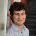 A young man with curly brown hair and a light blue shirt smiles while standing indoors near a doorway and a staircase.