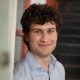 A young man with curly brown hair and a light blue shirt smiles while standing indoors near a doorway and a staircase.