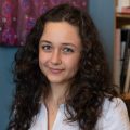 A young person with long curly hair wearing a white shirt stands indoors near a bookshelf and a patterned artwork on the wall.