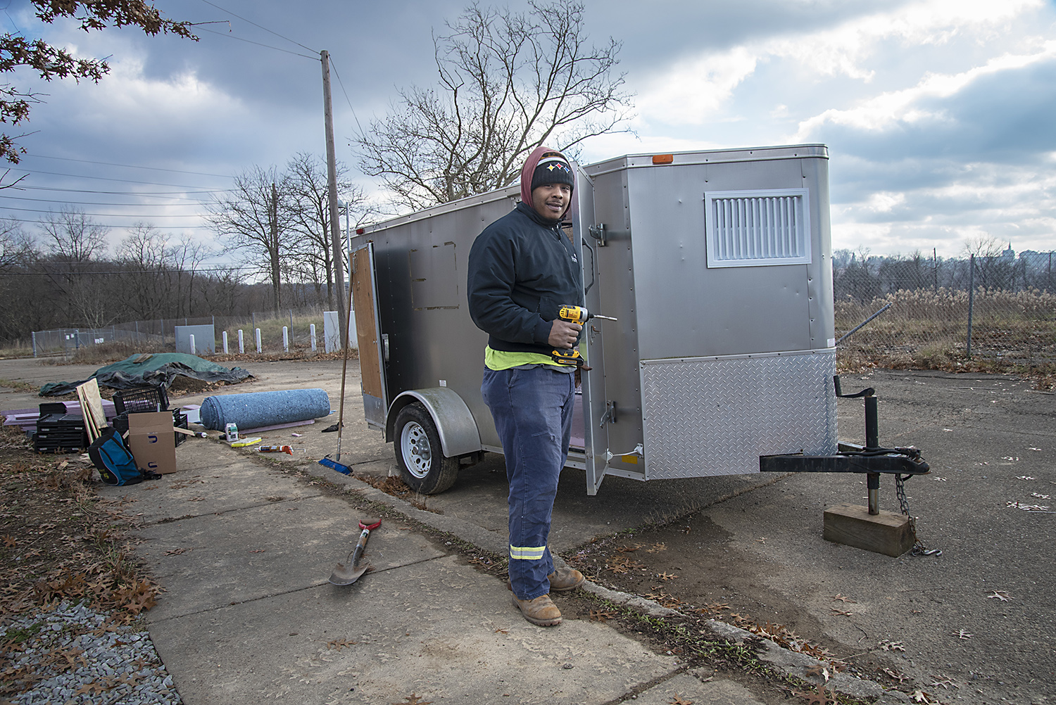 A person standing beside a silver enclosed trailer on a paved area, holding a drill with tools and materials scattered nearby on a cloudy day.