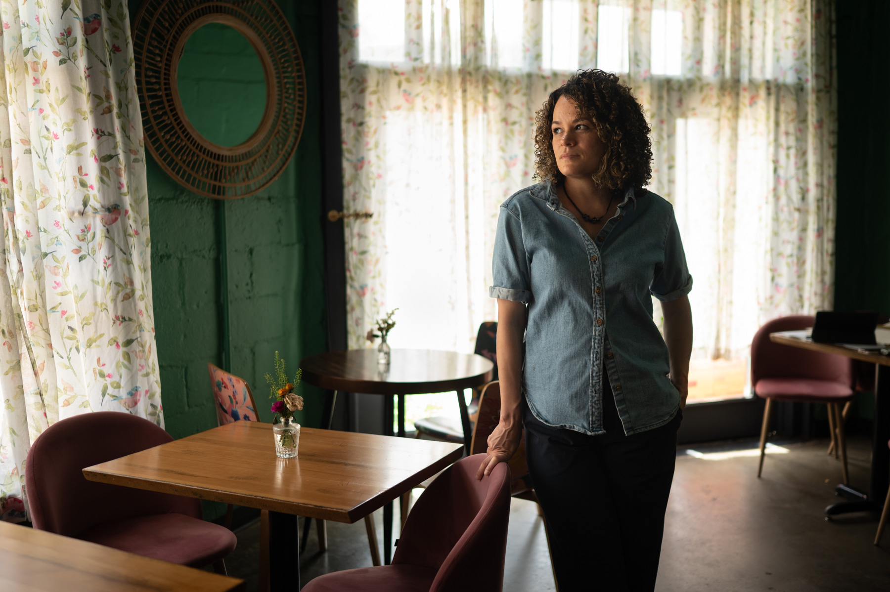 A person with curly hair in a denim shirt stands by a table in a sunlit café with pink chairs and floral curtains.