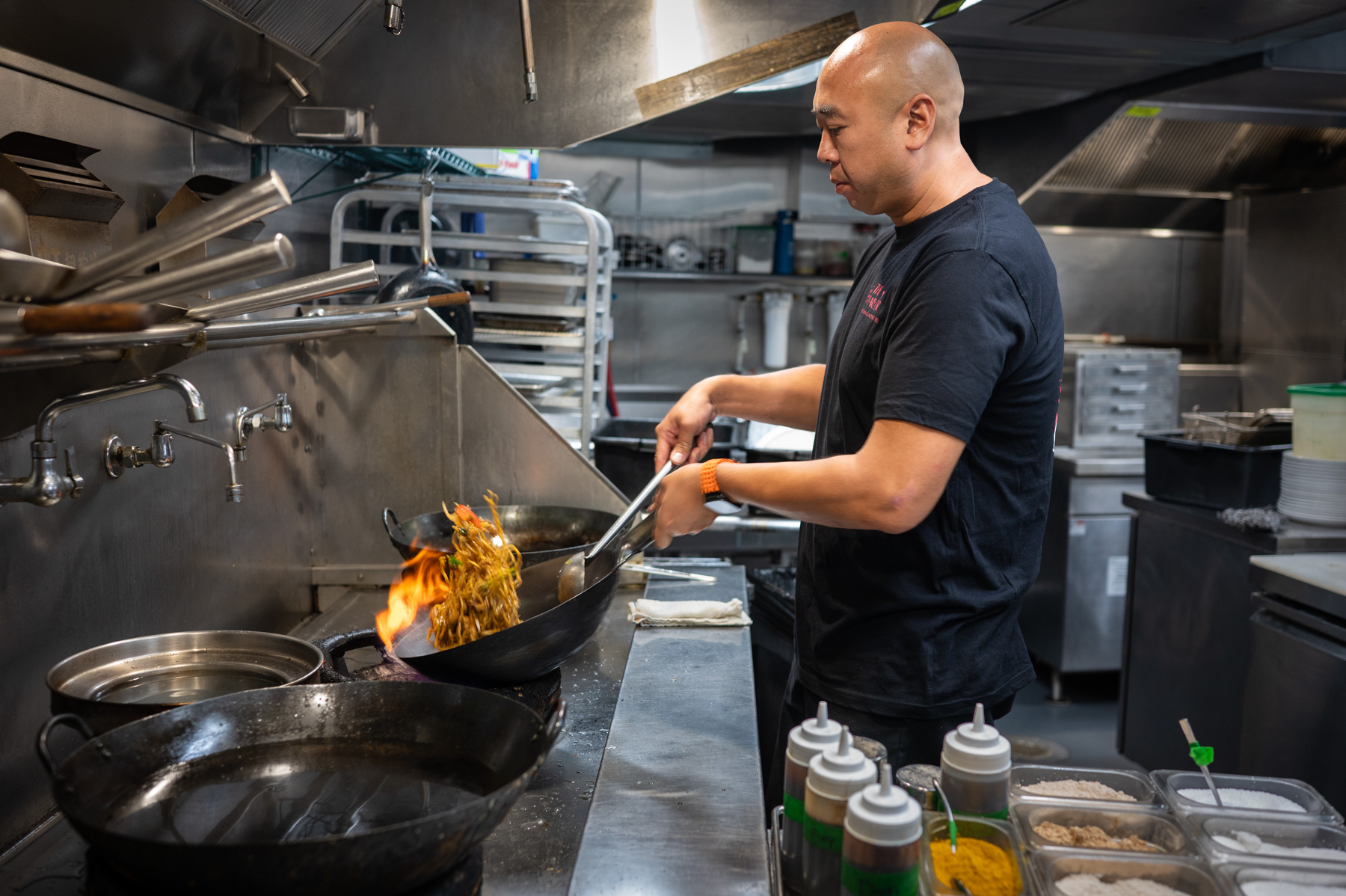 A chef in a commercial kitchen cooks noodles in a wok over an open flame, surrounded by cooking utensils and ingredients.
