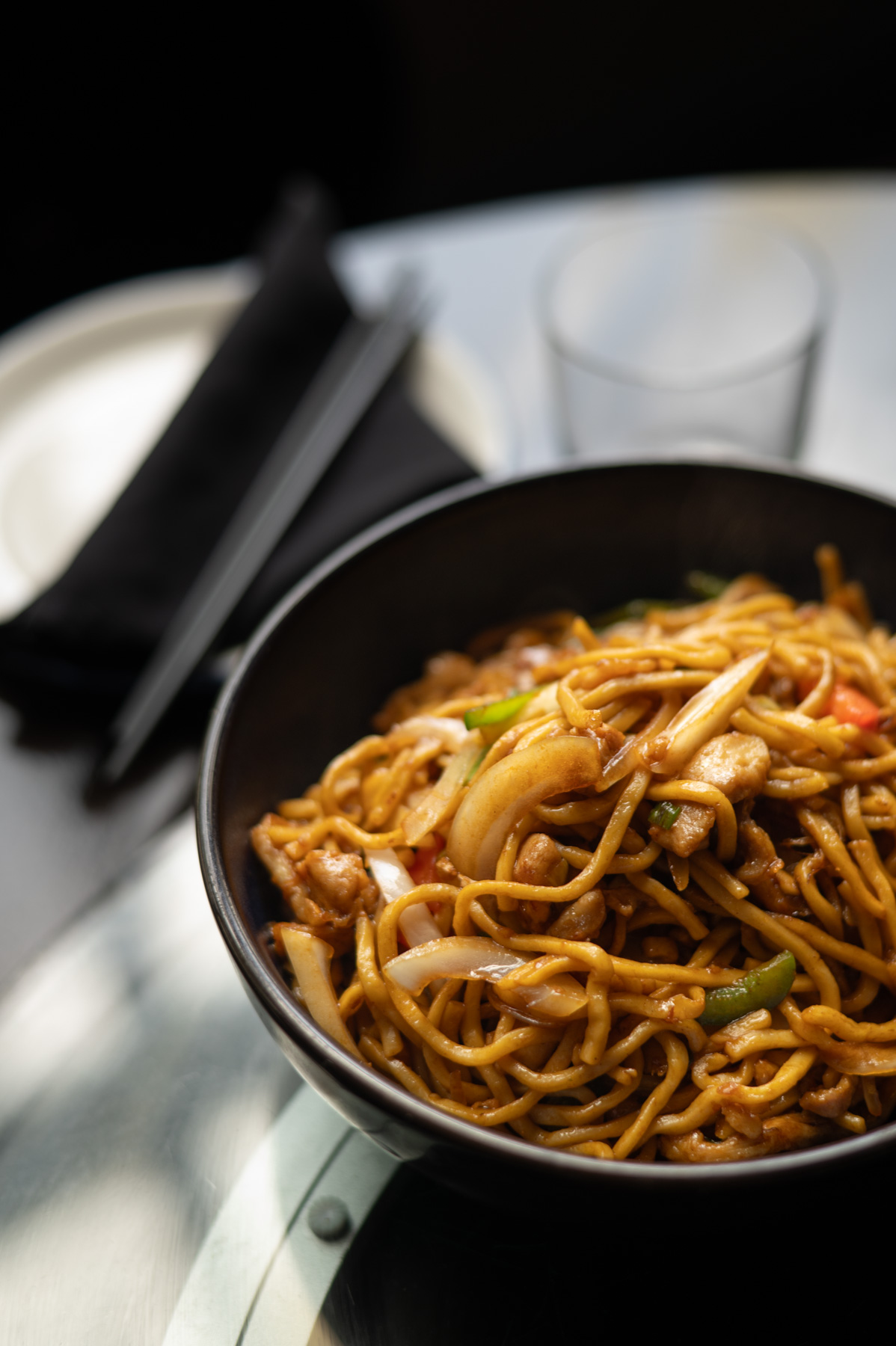 A black bowl filled with stir-fried noodles, chicken, onions, and vegetables sits on a table next to a glass, a plate, and chopsticks wrapped in a napkin.