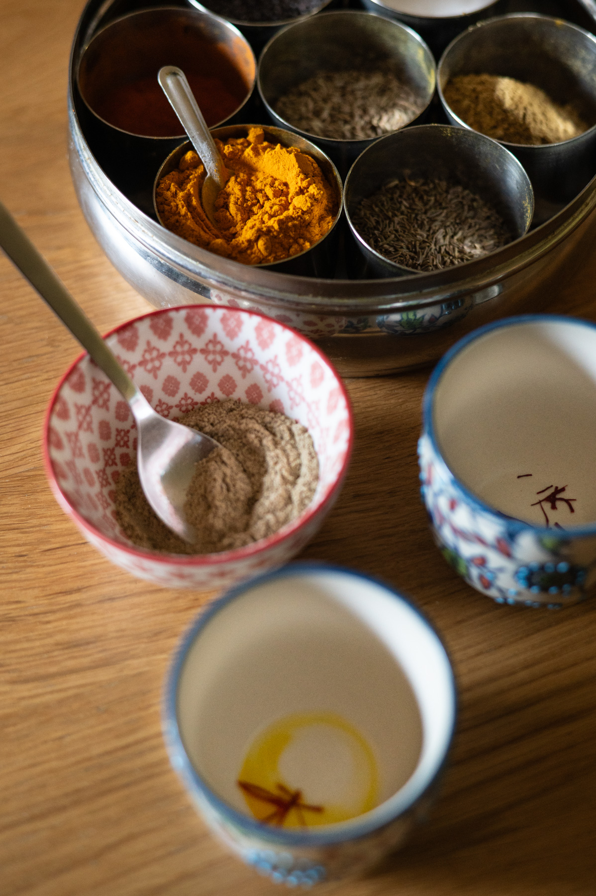 A spice box with various spices, a bowl with powder and a spoon, and two decorated cups, one with oil and a star anise, on a wooden surface.