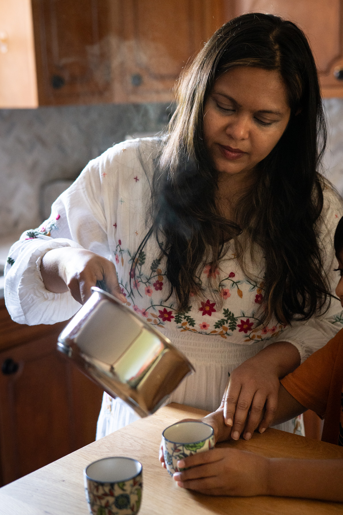 A woman in a white floral dress pours a hot beverage from a metal pot into a cup while a child sits beside her at a wooden table.