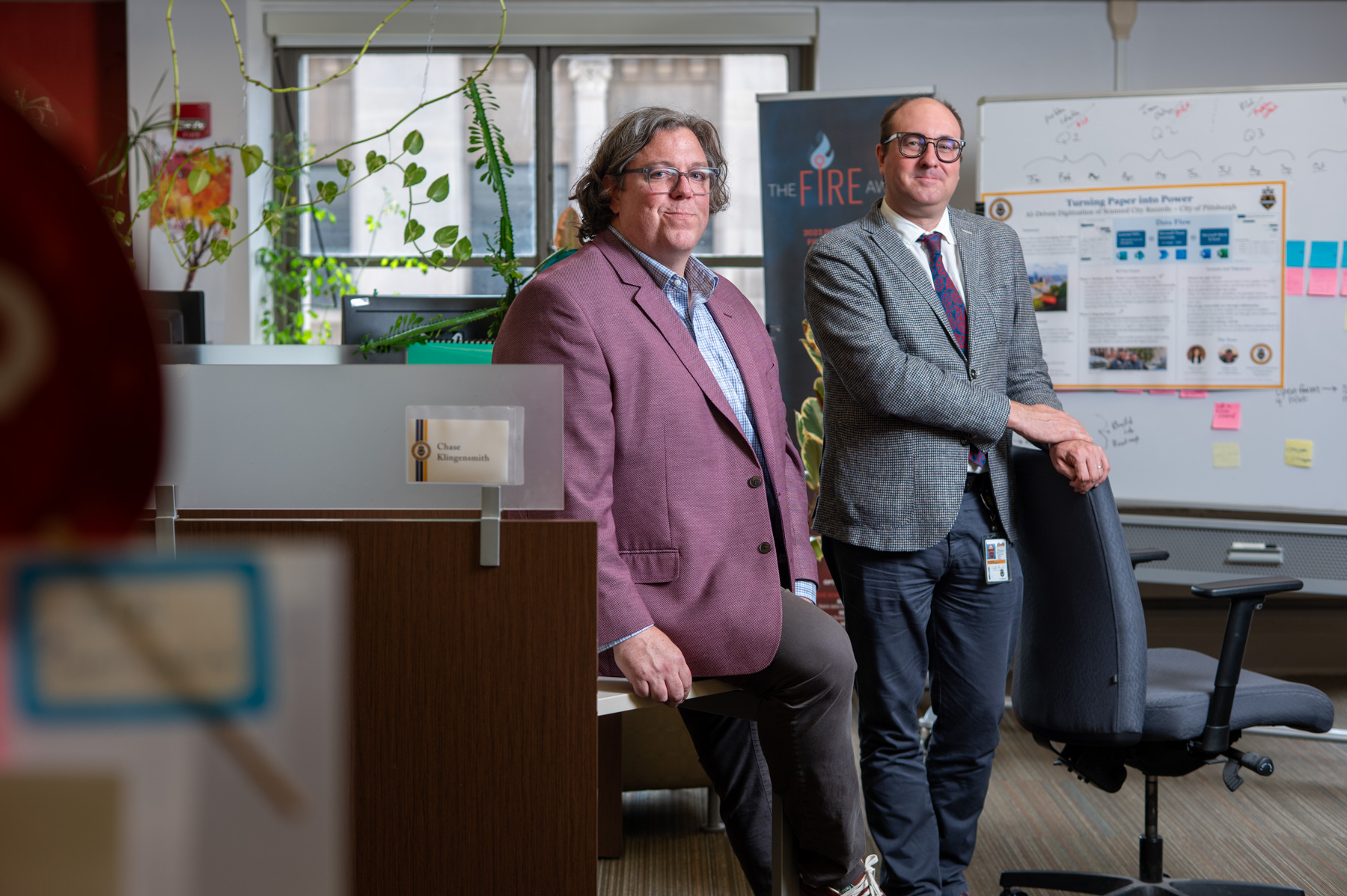 Two men in business attire pose in an office space with desks, whiteboards, a poster, and plants visible in the background.