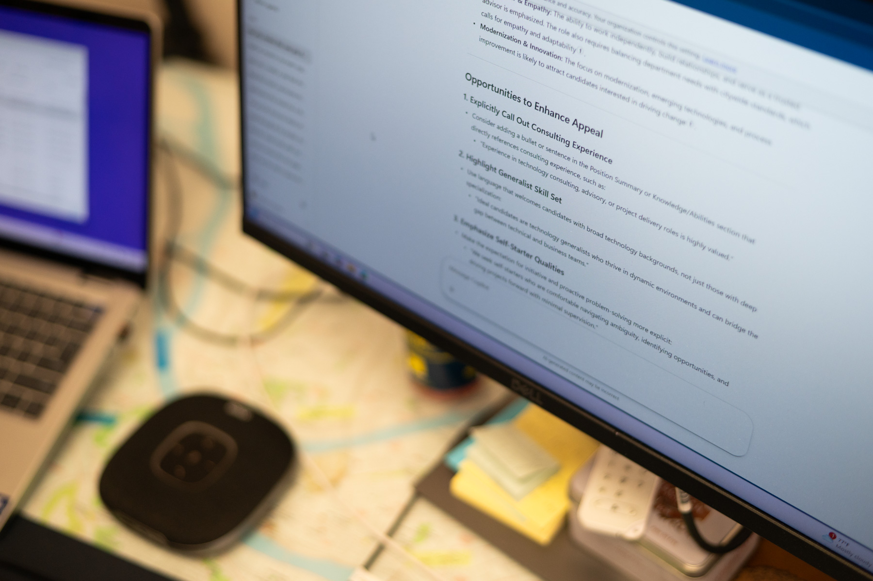 A computer monitor displays a text document next to a laptop and electronic devices on a cluttered desk with papers, cables, and sticky notes.