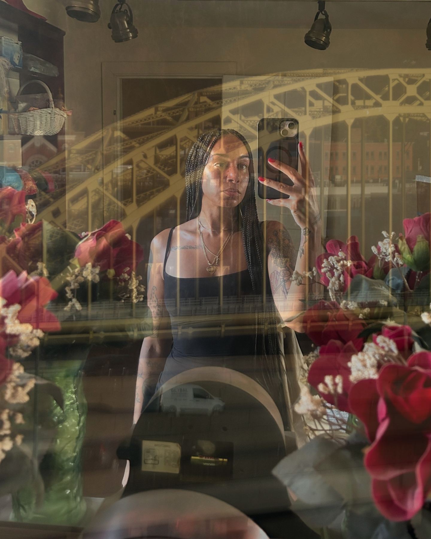 A person takes a mirror selfie surrounded by red flowers, with a bridge and city buildings reflected in the background.