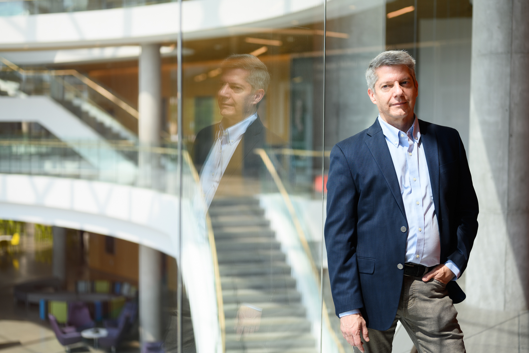 A man in a blazer stands by a glass wall inside a modern building, with his reflection visible and a staircase in the background.