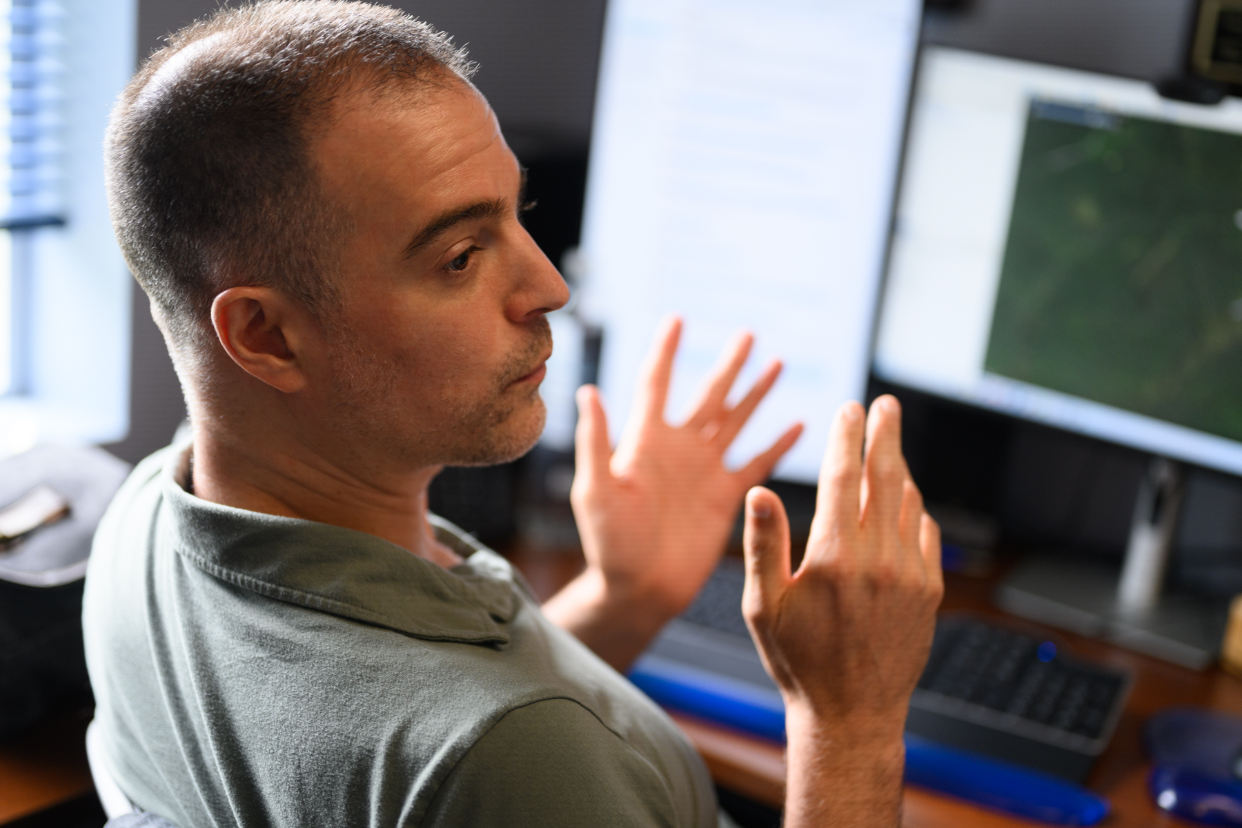 A person sits at a desk with multiple monitors, gesturing with both hands while speaking or explaining something.