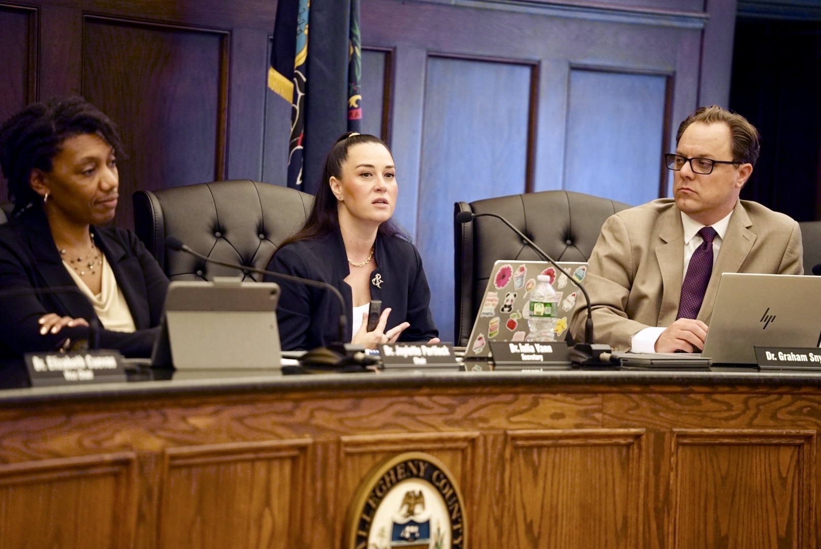 Three people sit at a panel discussion table with laptops and microphones, appearing engaged in conversation in a wood-paneled room with official emblems and flags in the background.