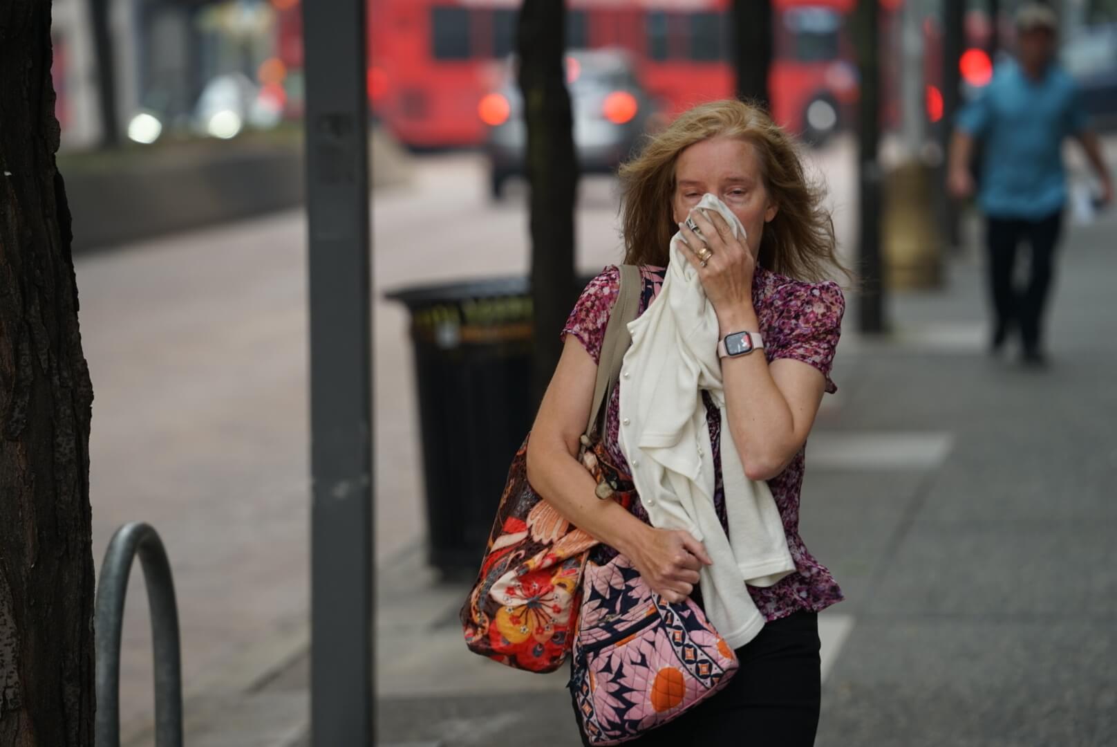 A woman walks on a city sidewalk holding a tissue to her nose and mouth, carrying a floral bag and a white cloth, with blurred people and vehicles in the background.