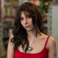 A woman with long brown hair wearing a red tank top and a heart-shaped necklace stands indoors, looking at the camera with a neutral expression. Bookshelves and plants are in the background.