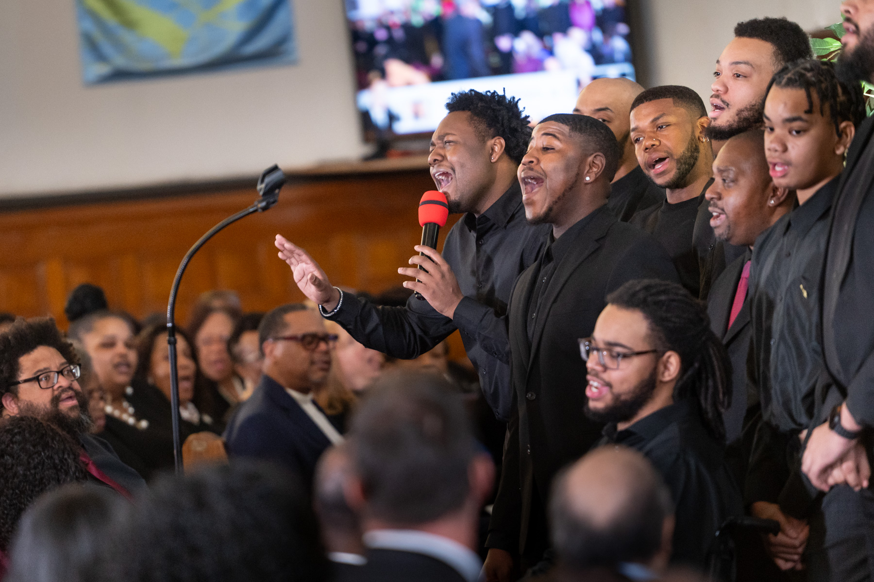 A group of men in black suits sing together in front of an audience in a wood-paneled room, with a TV screen and flag visible in the background.