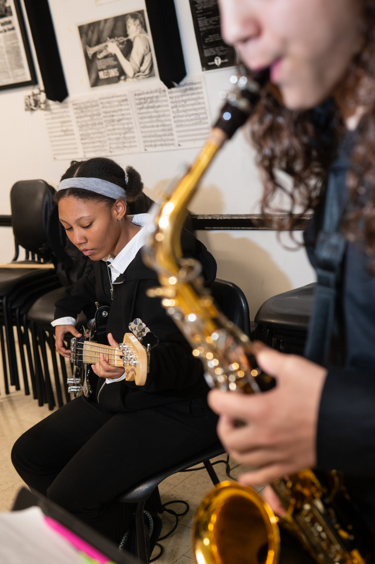 A student plays an electric bass guitar while another student plays a saxophone in a music classroom with chairs and sheet music in the background.