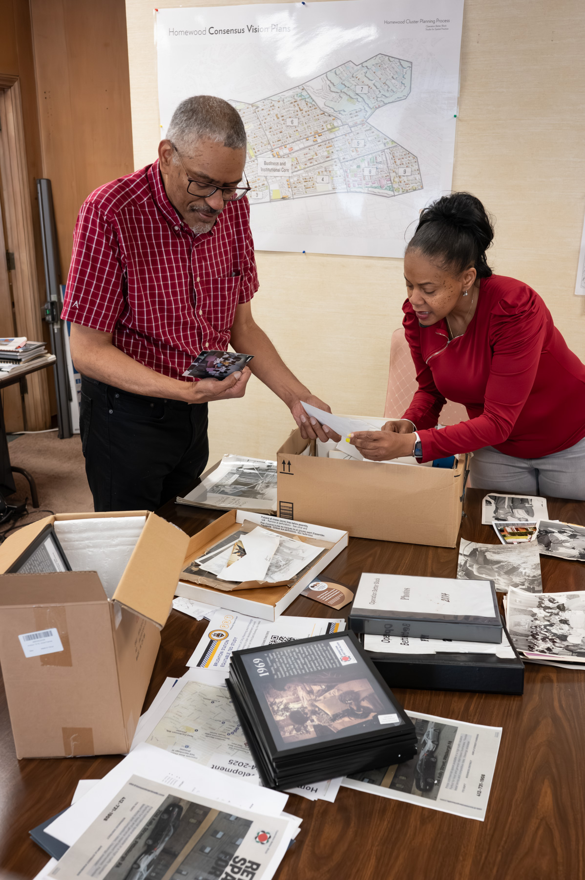 Two people review old photographs and documents on a table in an office, with boxes, binders, and a neighborhood map visible in the background.