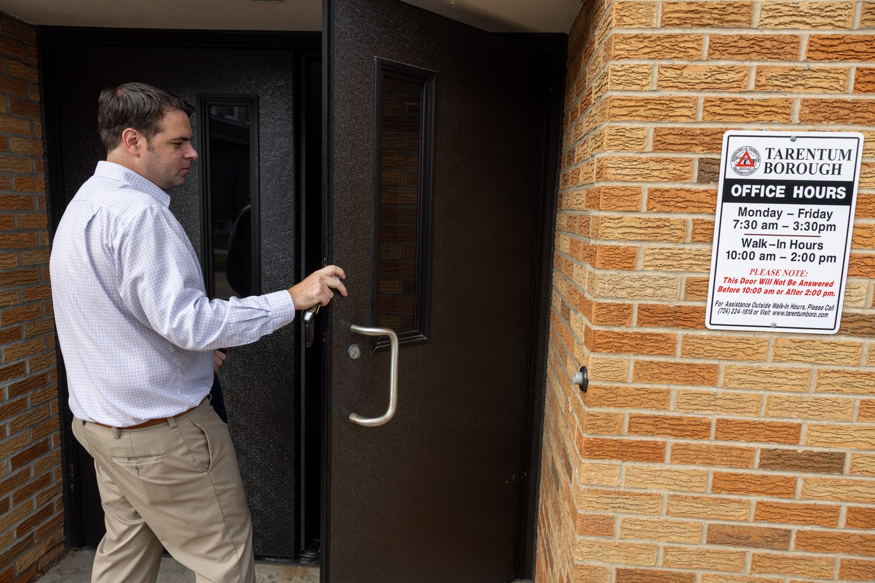 A man in a dress shirt opens a door next to a brick wall with a posted sign displaying Tarentum Borough office hours and visitor instructions.