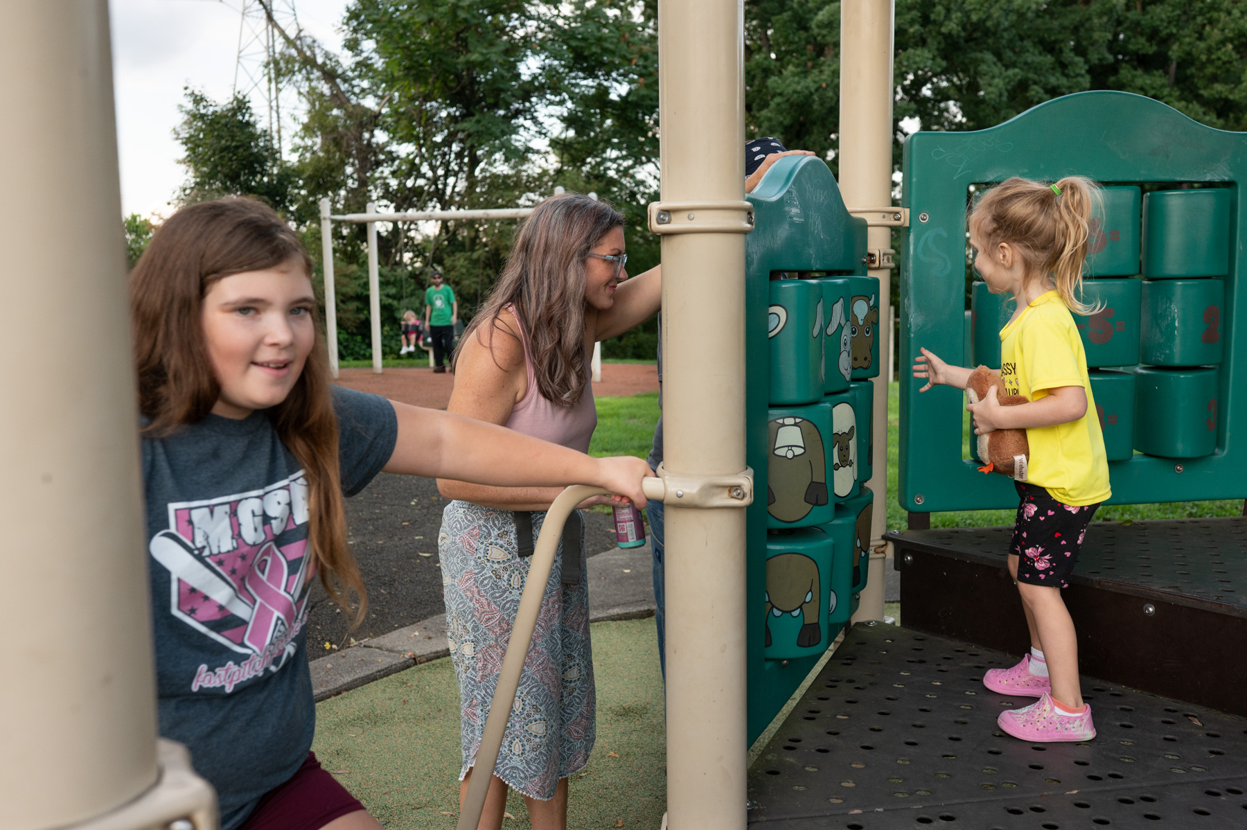 Three people, including two children and one adult, play on a playground climbing structure, with trees and grass visible in the background.