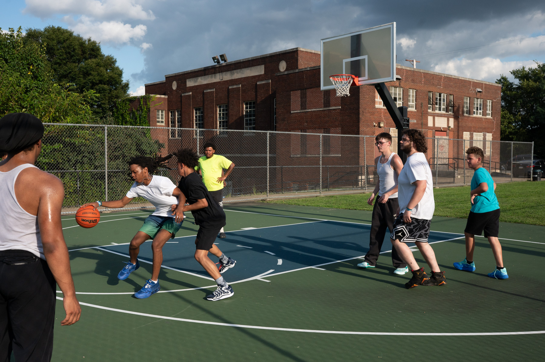 Six people play basketball on an outdoor court near a brick building, while another person stands on the sidelines observing the game.