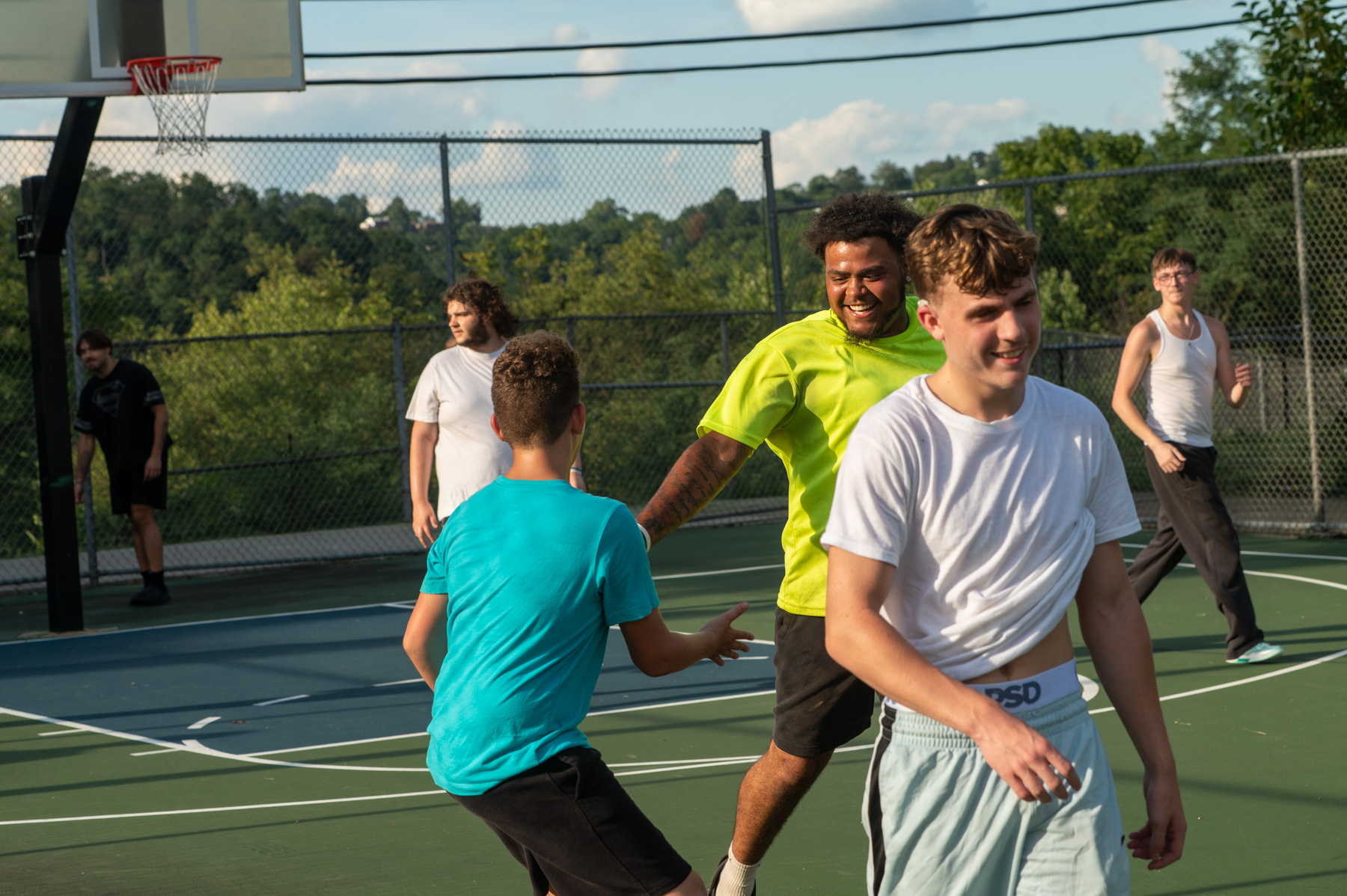 Six young men play basketball on an outdoor court under sunny weather, smiling and interacting as they move across the court. Trees and a fence are visible in the background.