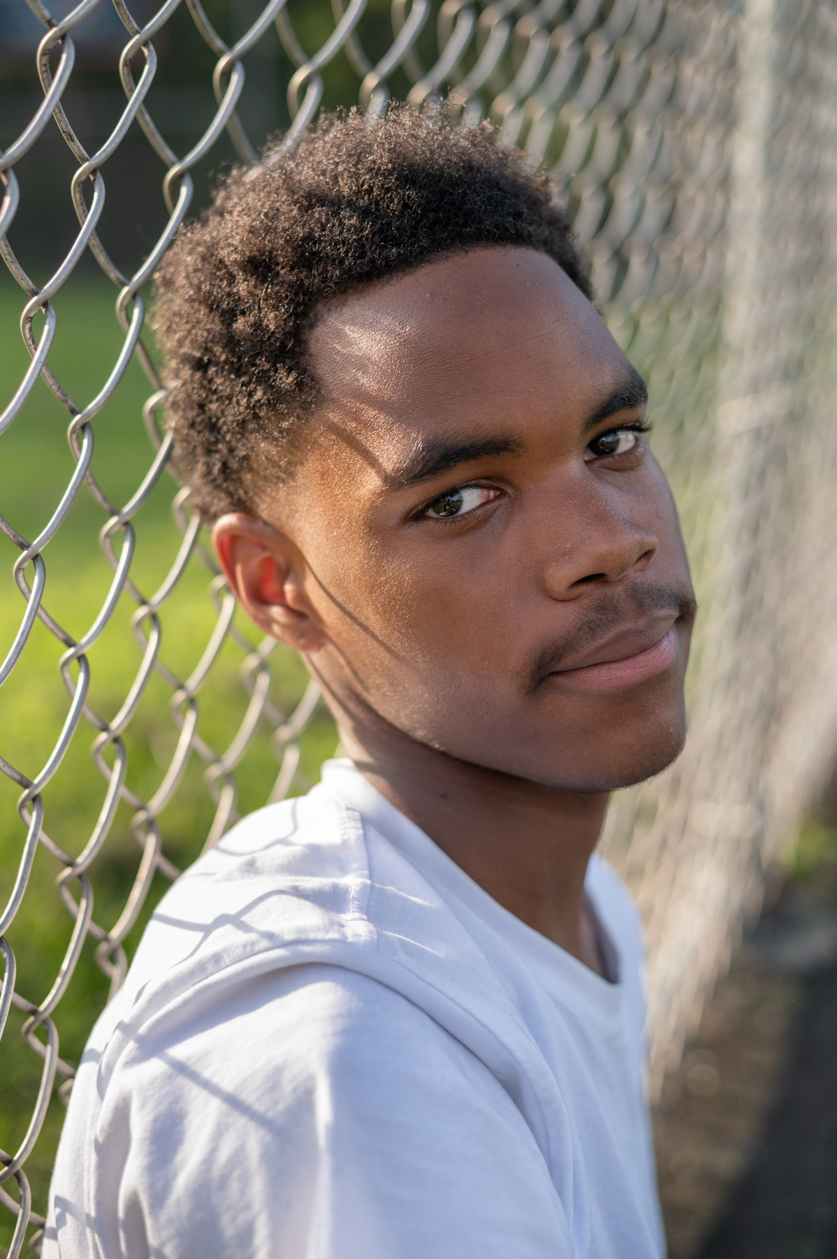 A young person in a white shirt sits by a chain-link fence, looking directly at the camera with a neutral expression.