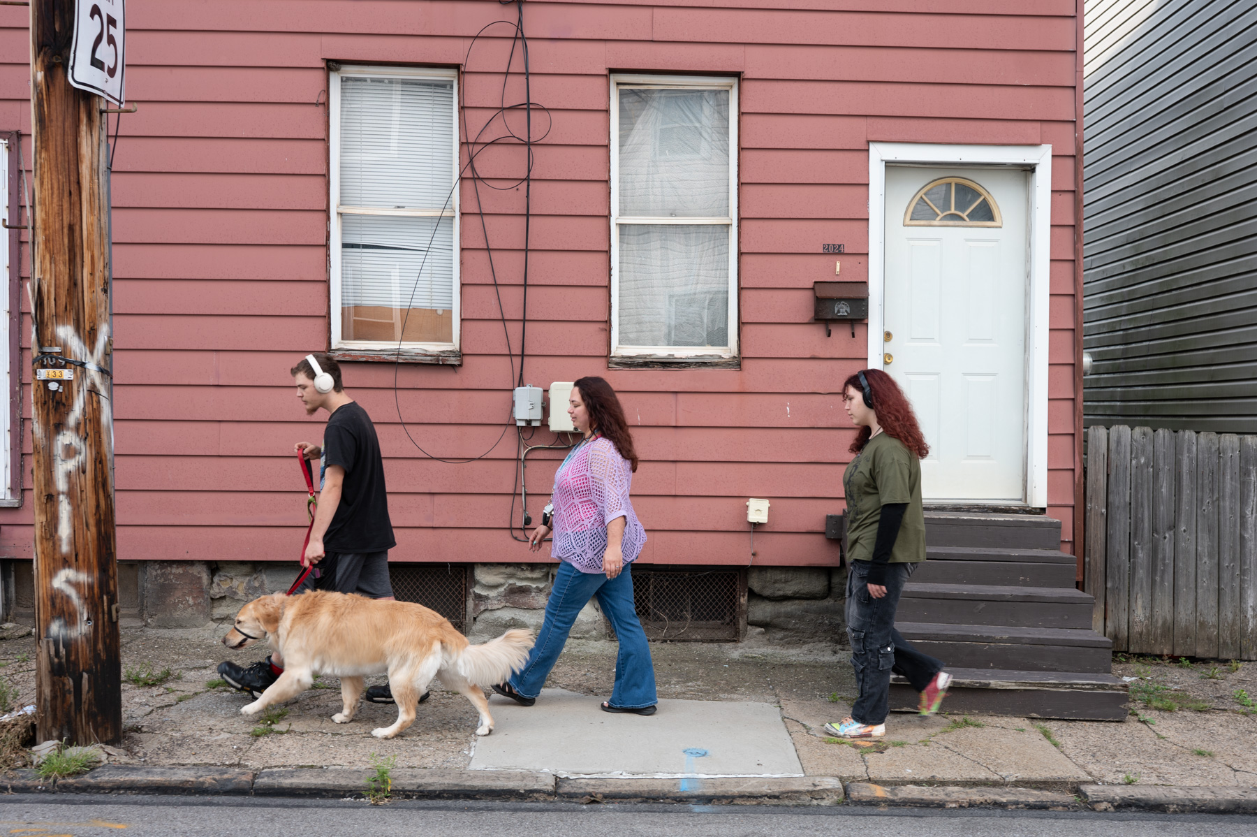 Three people, one wearing headphones and walking a dog, pass a pink house with a white door and windows on a sidewalk.