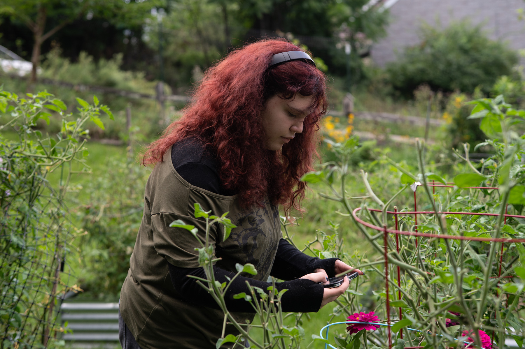 A person with long red hair uses a smartphone while standing among green plants and flowers in an outdoor garden.