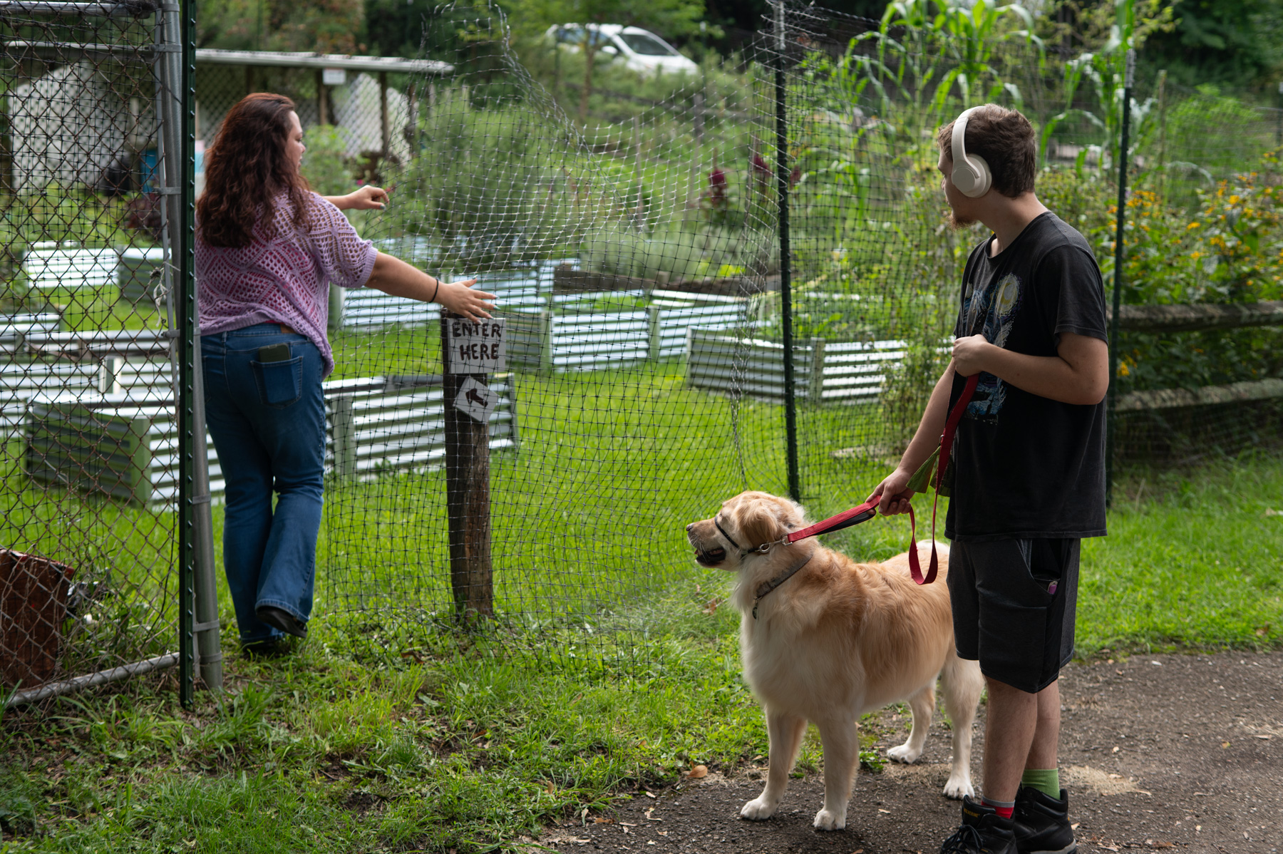 A woman points to an “Enter Here” sign on a garden fence while a boy with headphones holds a golden retriever on a leash nearby.