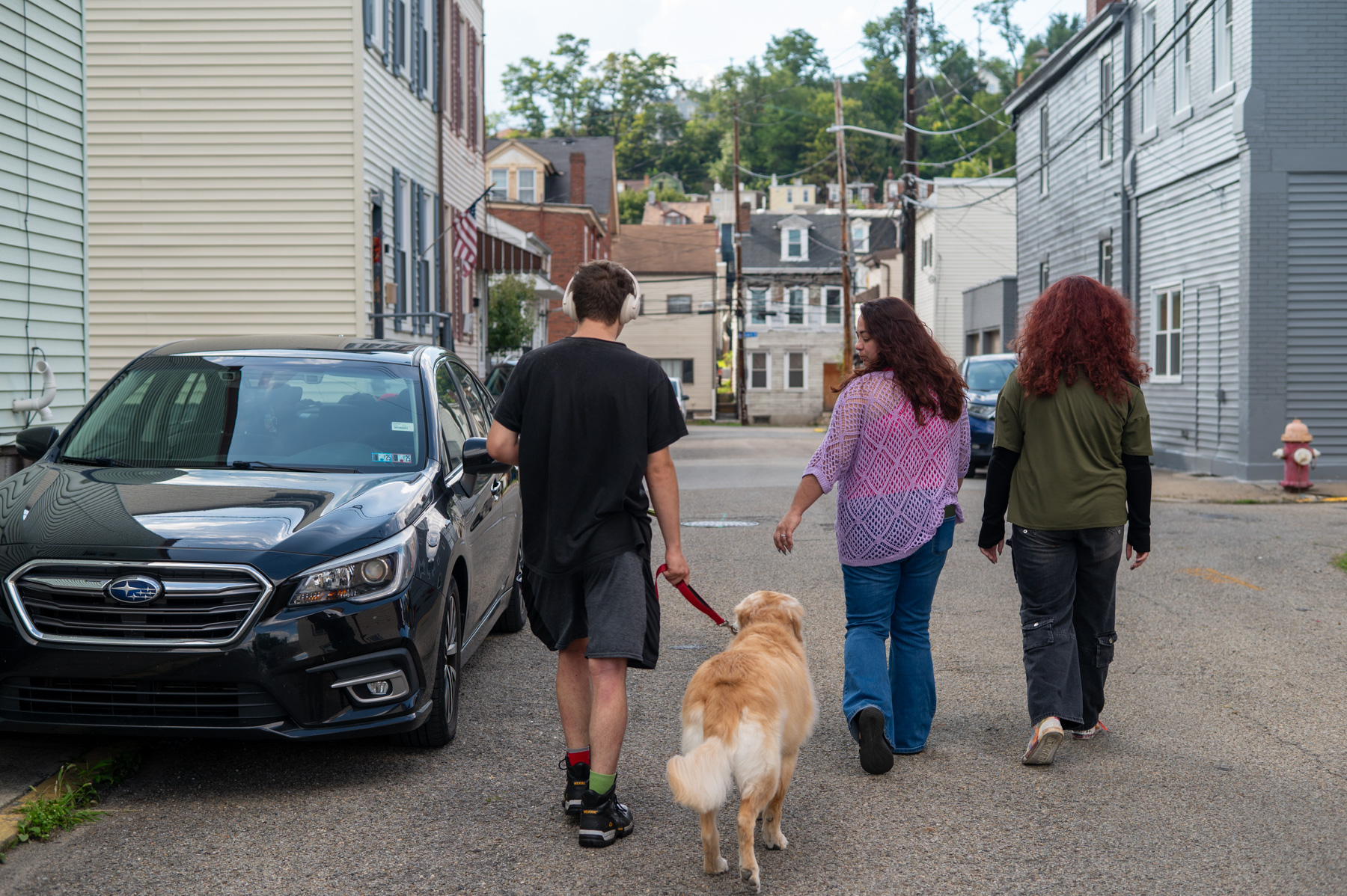 Three people walk down a residential street with a golden retriever on a leash. Parked cars and houses line both sides of the street.