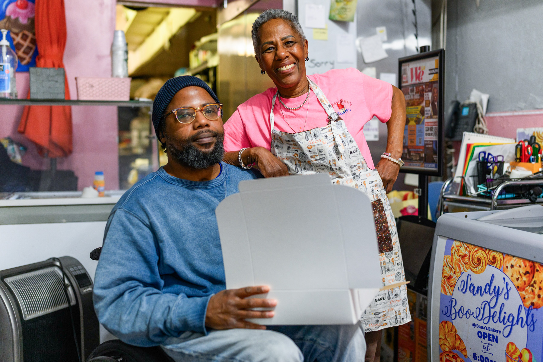 A man in a wheelchair holds an open box while a woman in an apron stands beside him, both smiling, inside a bakery or food shop.