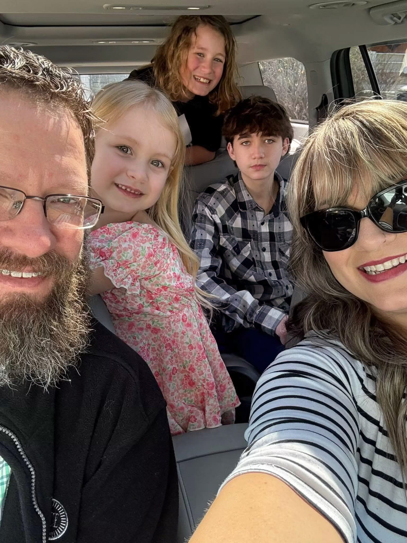 A group of five people, including two adults and three children, sit inside a car, smiling and posing for a selfie in daylight.