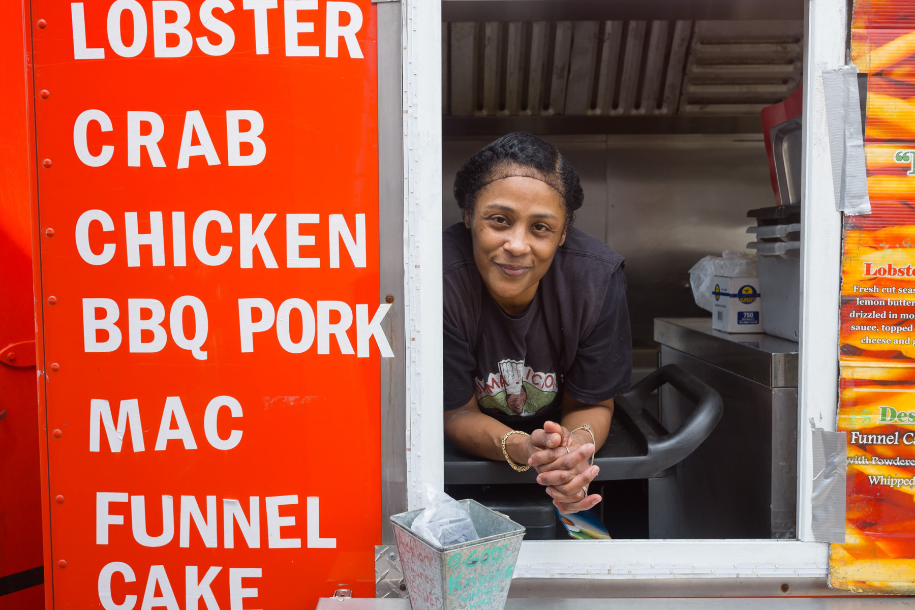 A woman leans out of a food truck window next to a menu listing items like lobster, crab, chicken, BBQ pork, mac, and funnel cake.