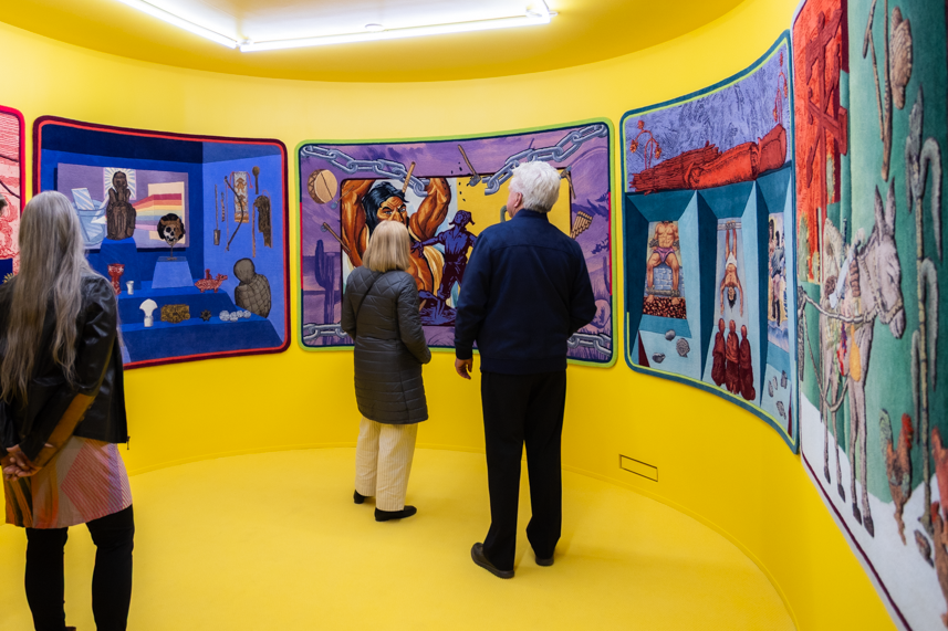 People look at brightly colored paintings in a bright yellow painted room with curved walls.