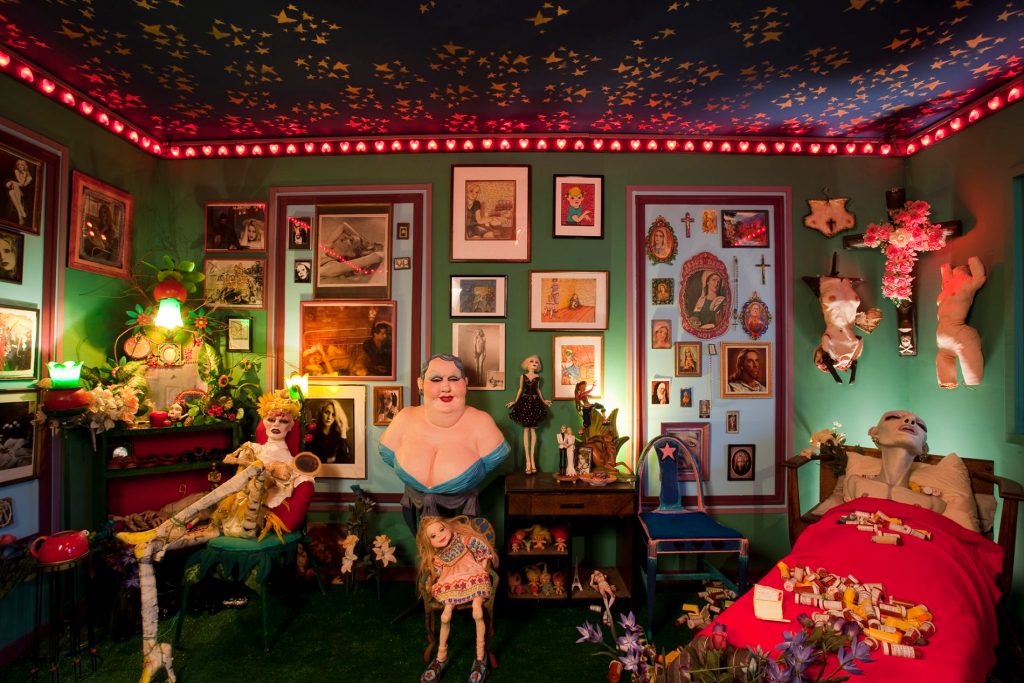 A green room with many pieces of art on the wall, with a skeleton, a bust of a person in a dress, and an emaciated person in a bed prominently featured.